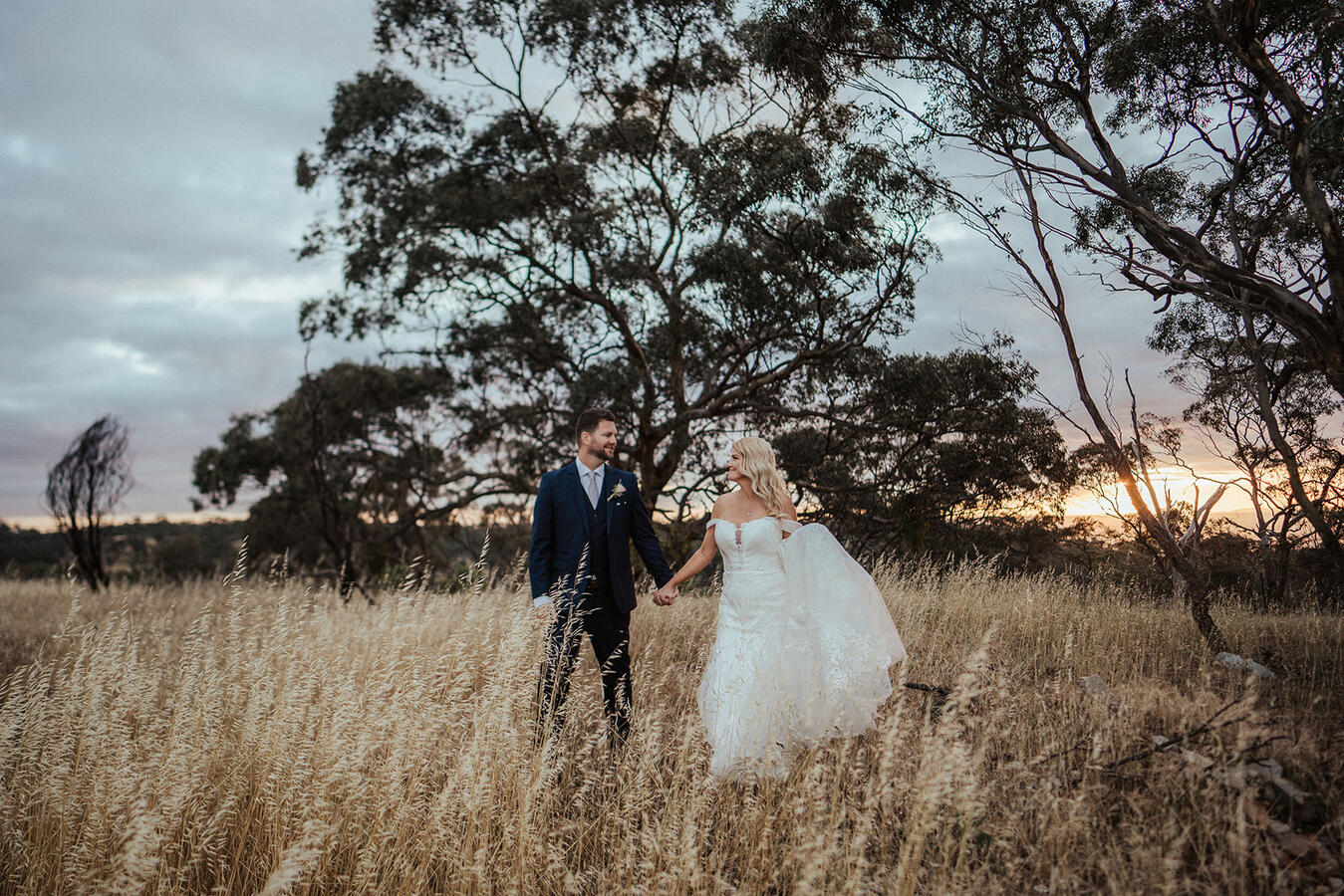 A bride and groom holding hands in a field of tall grass at sunset.
