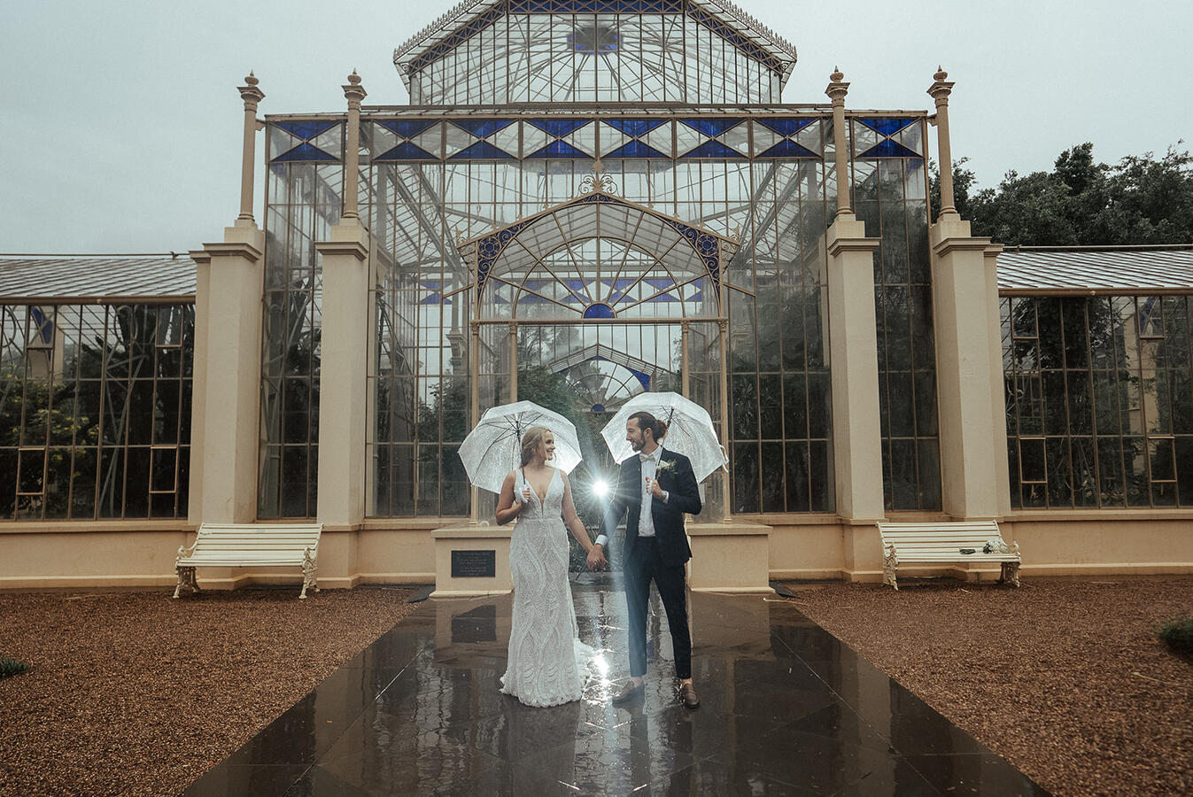 A bride and groom holding hands in the rain in front of a glass house at the bontanic gardens in Adelaides CBD