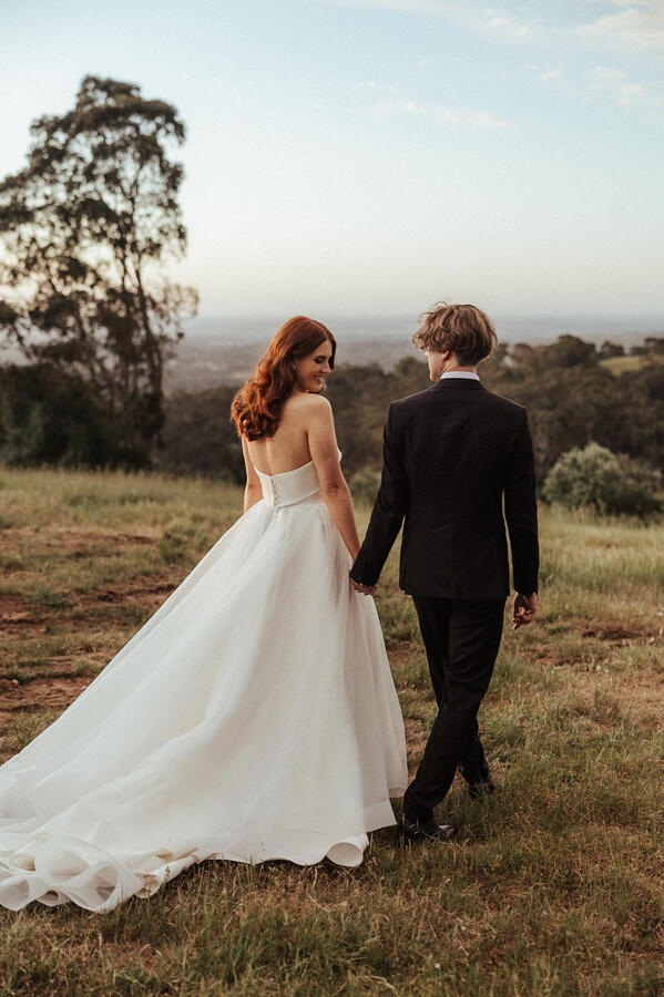A bride and groom walking in a field at sunset, smiling at each other.